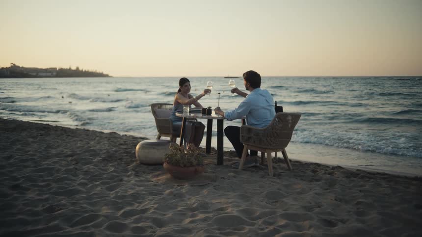 A couple sits at a small table on a sandy beach and toasts with wine glasses as waves break on the shore during a golden sunset
