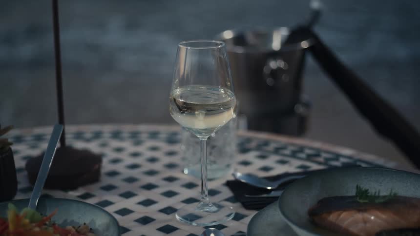 A man in a light blue shirt sips wine at the beach as twilight sets in