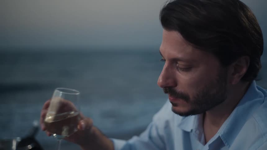 A man in a light blue shirt enjoys a glass of white wine by the ocean at dusk