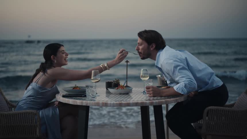 A couple enjoys a romantic seaside dinner at twilight with wine and gourmet dishes by the shore