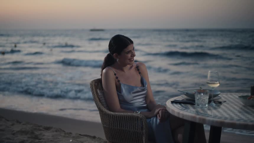 Woman in a silver dress sits at a seaside table at sunset