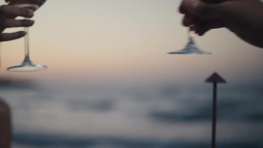 Two hands toast with glasses of white wine against the backdrop of a calm dusky ocean