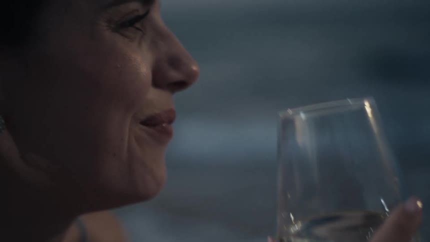 The video shows a closeup of a woman at a table drinking from a wine glass she takes a sip and as she lowers the glass a smile spreads across her face