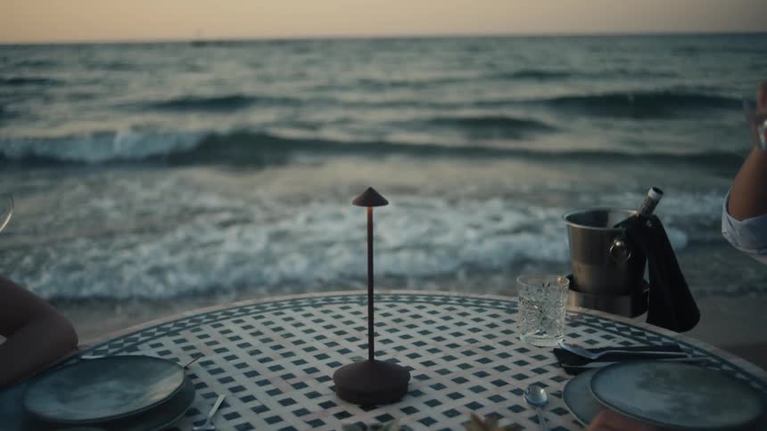 The video shows a romantic toast with two people raising glasses of white wine the camera then pans down to show the waves of the ocean gently lapping at the shore in the background