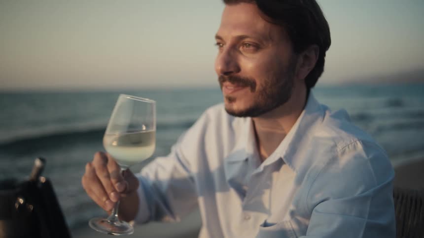 The video shows a man sitting on a beach at what appears to be sunset he is holding a glass of white wine takes a sip and then smiles as he looks out at the ocean