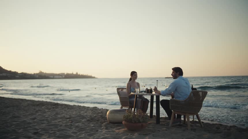 The video shows a romantic dinner scene on a beach at sunset a man and a woman are seated at a small table with the waves crashing gently in the background