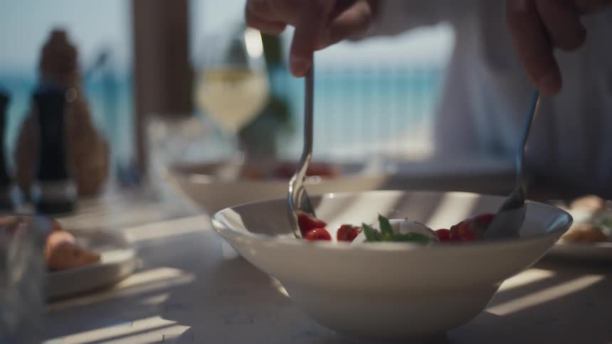 A hand with a spoon serves a salad of mozzarella tomatoes and green leaves from a white bowl on a table with a blurry ocean and wine glass visible in the background