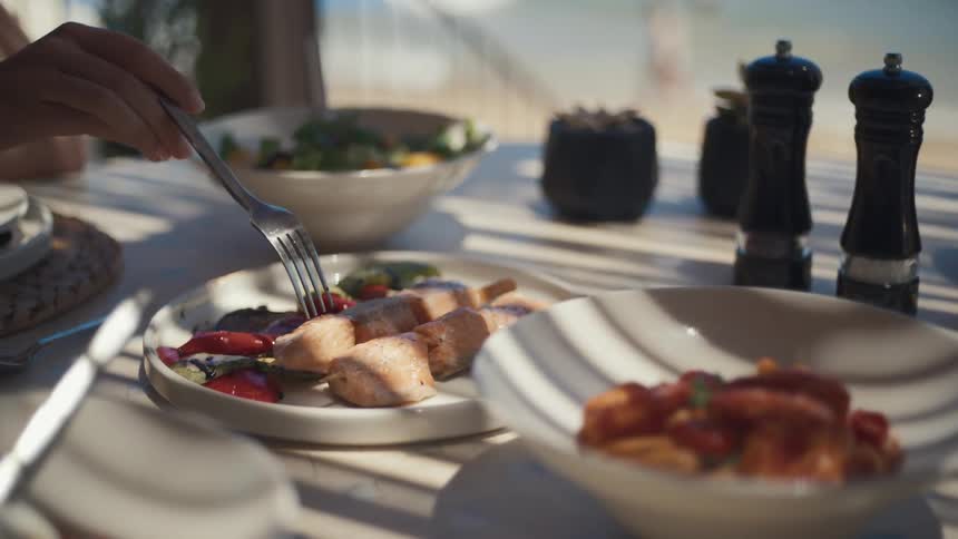 Hand holding a fork over a plate of grilled salmon with vegetables accompanied by bowls of salad and gnocchi
