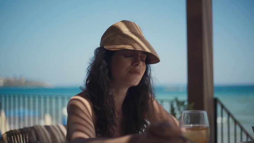 The video shows a woman with long dark hair and a straw bucket hat sitting at an outdoor table she is eating a meal and has a glass of wine in front of her with a view of the ocean in the background