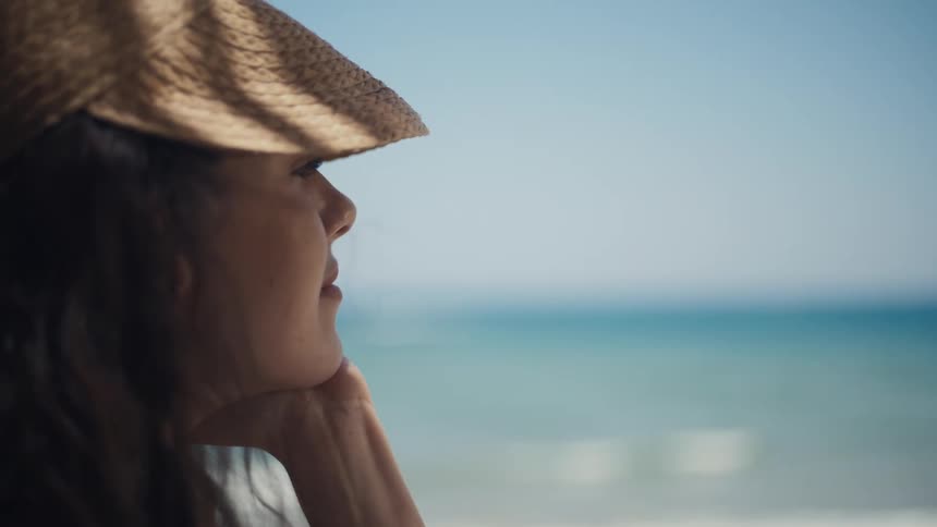 A woman in a widebrimmed straw hat gazes out at the sea on a bright sunny day