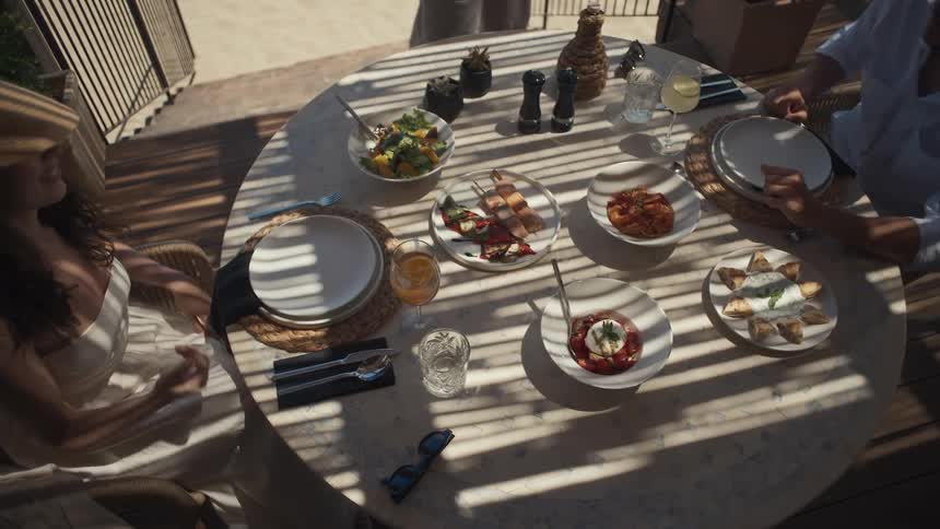 A highangle shot shows a group of people enjoying an outdoor lunch together with various plates of food spread across a round table as they raise their glasses for a toast