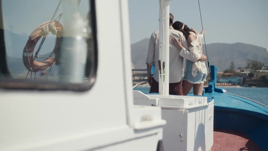 A couple stands on the deck of a boat with the mans arms around the woman as she rests her head on his shoulder all while looking out at a coastline with a mountain in the distance