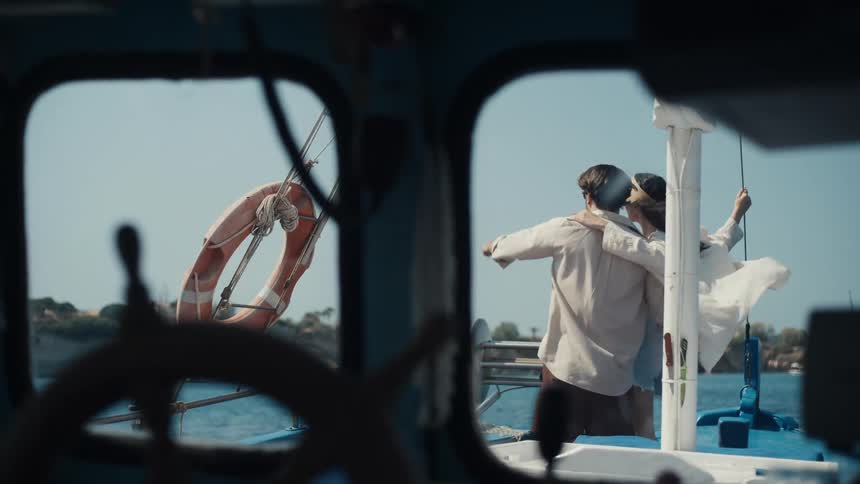 Framed by a boats window a couple stands arm in arm on deck gazing out at the sea
