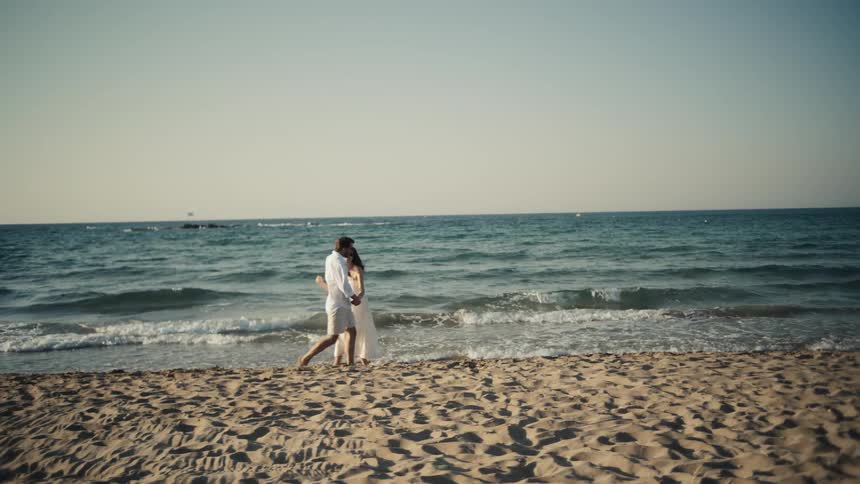 The video shows a couple holding hands and walking on a sandy beach they are both dressed in white with the woman wearing a dress and the man wearing a shirt and shorts