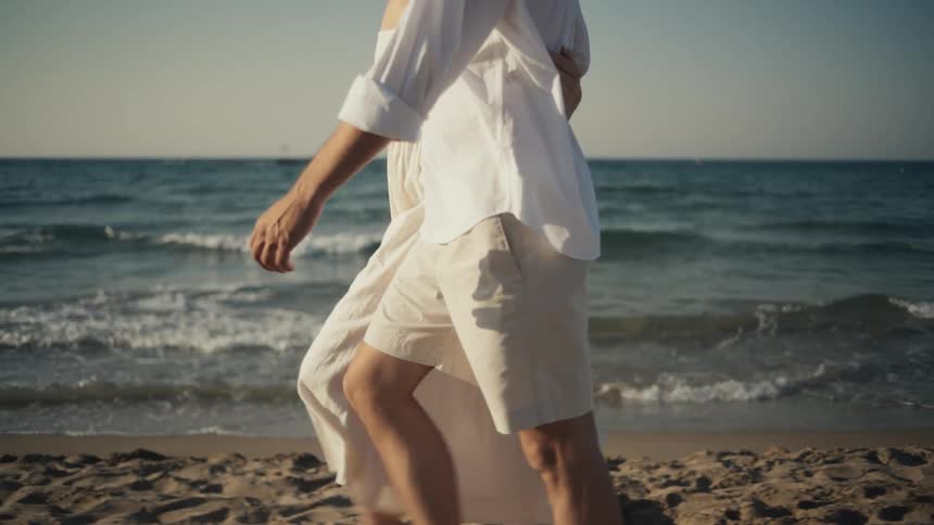 A couple walks on the beach holding each other wearing white clothes as the ocean waves hit the shore at midday