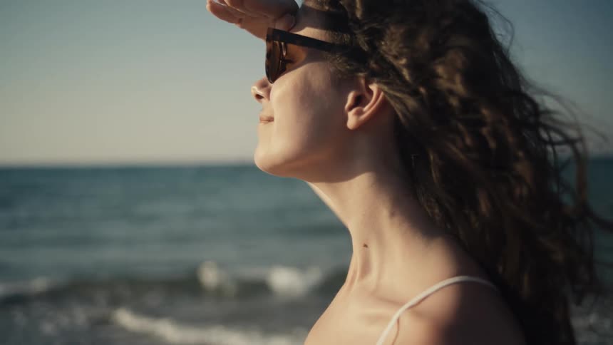 The video shows a closeup of a woman on a beach looking out at the ocean she is wearing sunglasses and shades her eyes with her hand as if looking into the sun her hair blows in the wind and she smiles as she lowers her hand