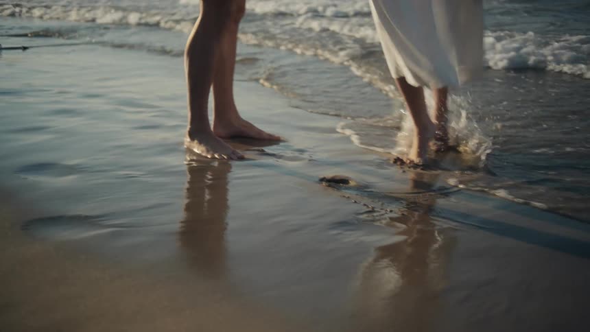 A man and a woman dance close together on a sandy beach as waves wash over their bare feet in the golden light
