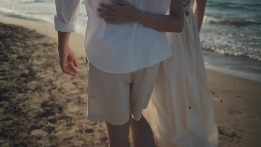 A couple walks barefoot along a sunlit sandy beach with the mans arm around the womans waist as they both look out at the waves