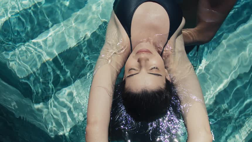 A woman in a dark swimsuit floats on her back in a bright blue swimming pool with her eyes closed and her hair floating around her head while being held by another person