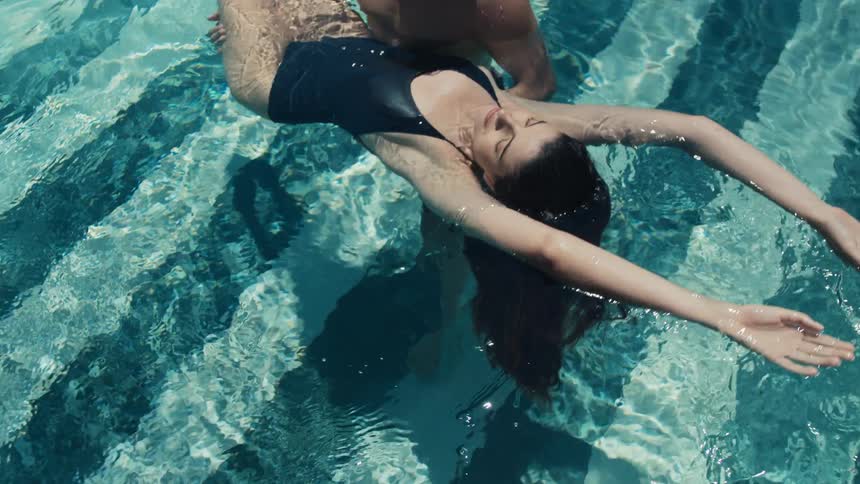 Woman in dark swimsuit floats on her back in pool with arms outstretched and held by a man