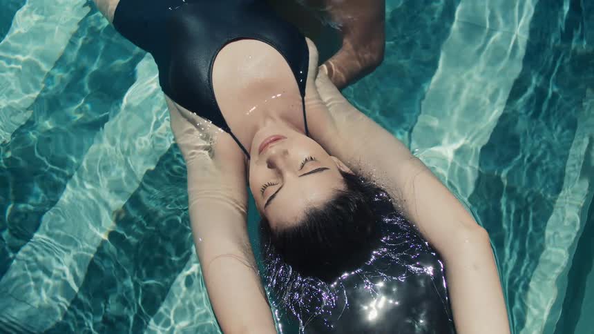 The video shows a highangle shot of a woman floating on her back in a swimming pool she is wearing a black onepiece swimsuit and has her eyes closed as her hair floats around her head in the water