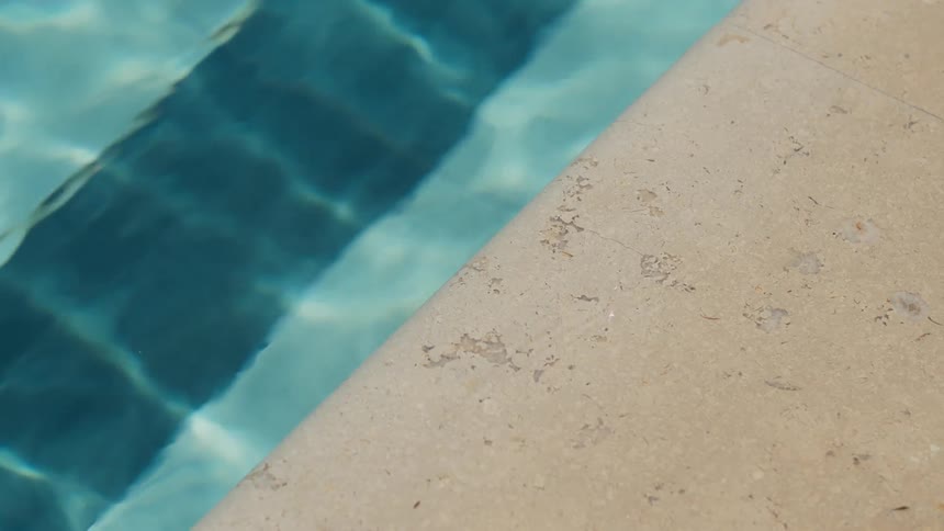The video shows a closeup of the edge of a swimming pool the left side shows the clear blue water with sunlight reflecting off the ripples while the right side shows the beige textured concrete of the pool deck