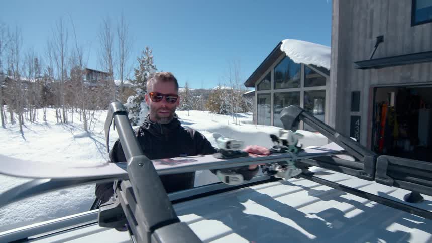 The video shows a man putting a snowboard and skis onto a roof rack on a car the scene is set in a snowy landscape in front of a modern home indicating a day of winter sports