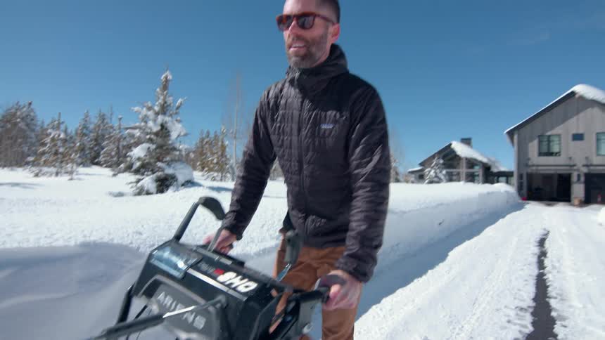 A man wearing sunglasses rolls a snow plow along his driveway in a suburban area at midday