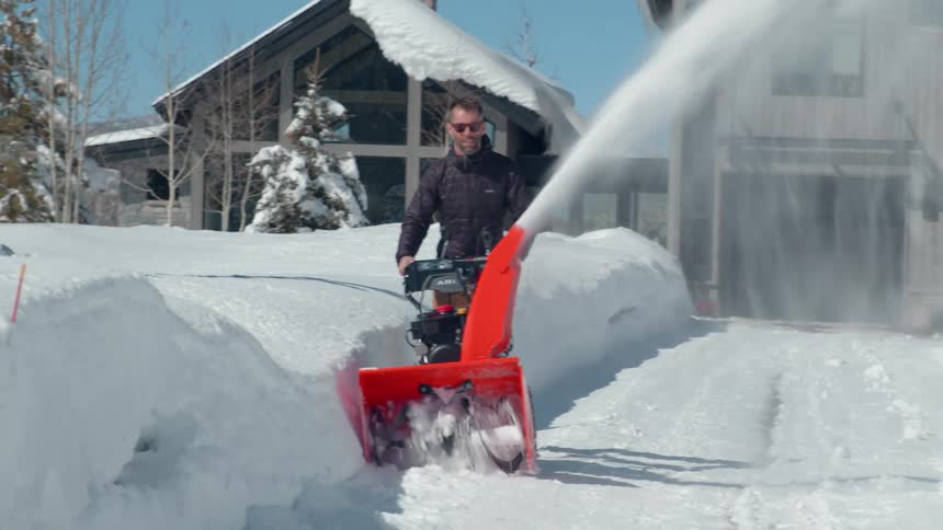 The video shows a man using a snow blower to clear a path in front of a house the machine is throwing a large stream of snow to the side creating a clear walkway