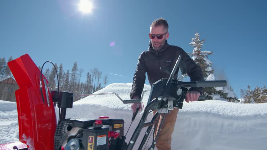 A man outside is starting a snowblower while surrounded by a lot of snow