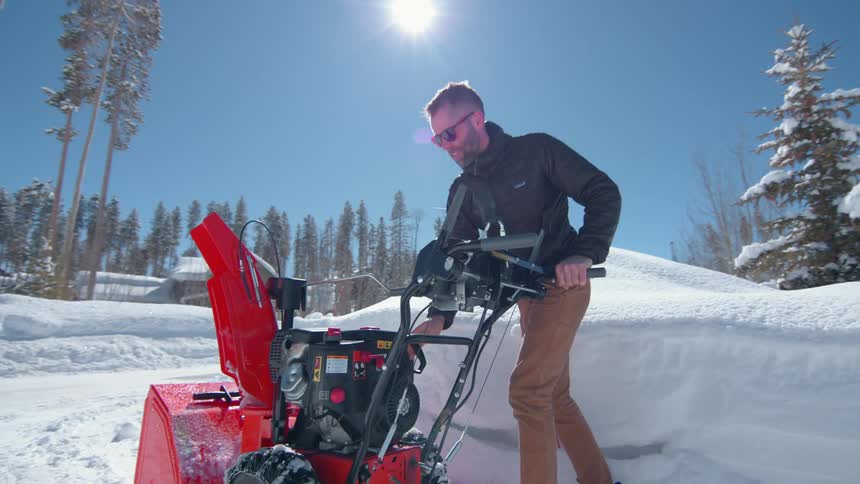 A man wearing a dark jacket and sunglasses is shown outdoors on a sunny day standing with a bright red snow blower and preparing to use it to clear snow