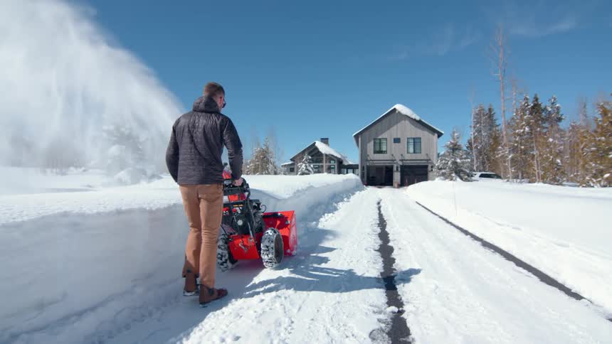A man wearing a gray jacket and tan pants uses a bright orange snowblower to clear a path on a snow covered driveway in front of two houses under a bright blue sky