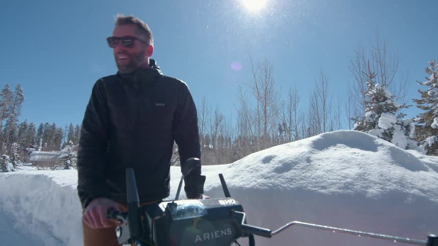 The video shows a man standing in a snowy landscape talking to a snow blower he is pushing he is wearing sunglasses and a black jacket and the sun is shining brightly creating lens flare