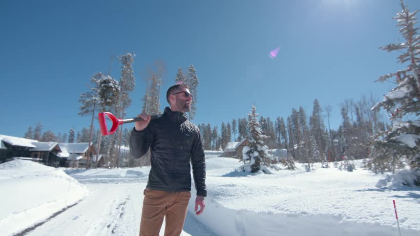 A man in sunglasses stands confidently holding a shovel in a snowy neighborhood resting a red shovel on his shoulder under a bright sky