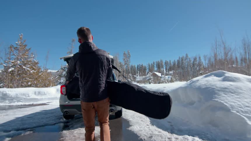 A man walks out of a garage and places a guitar in the trunk of a car with the street covered in snow during a winter midday
