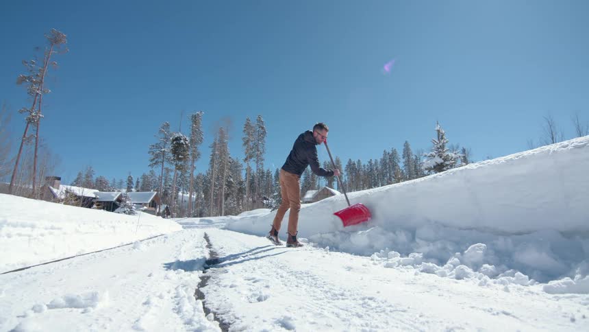 A man is seen from a low angle on a bright sunny day actively shoveling a large pile of snow from the side of a road in a wintry rural setting