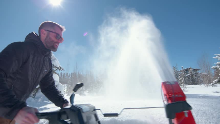 A man wearing sunglasses operating a snow blower clearing snow off the road at midday under the sun