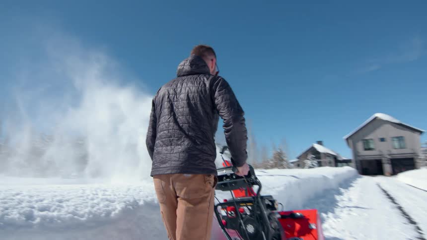 Man using a snow blower to clear the driveway in front of a house