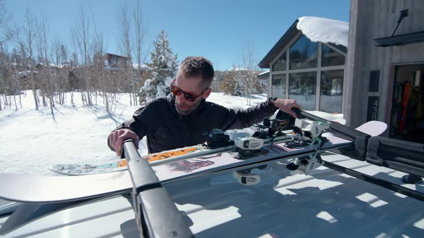 A man in a black jacket and sunglasses secures a pair of skis and a snowboard to a cars roof rack in front of a modern house on a snowy day