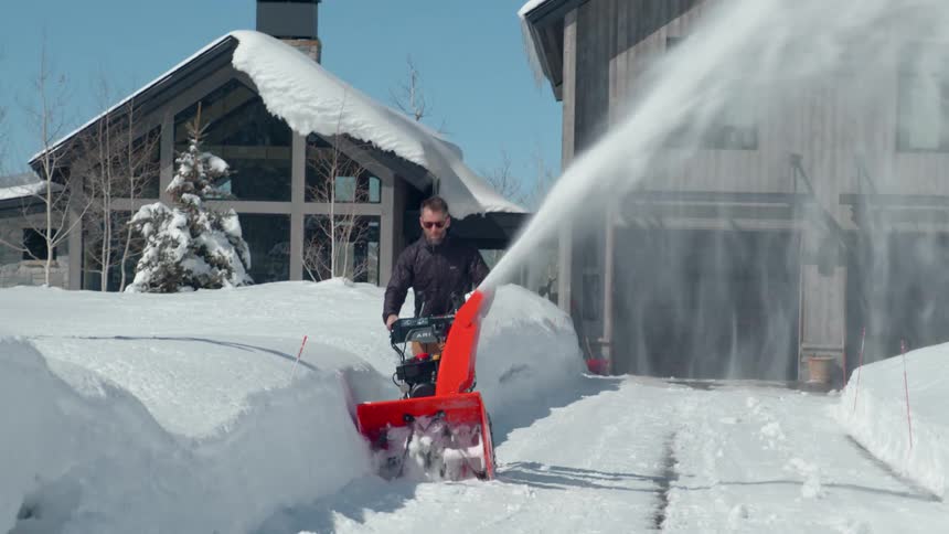 The video shows a man using a snow blower to clear a path in front of a rusticstyle house the snow is thick on the ground and rooftops indicating a heavy snowfall