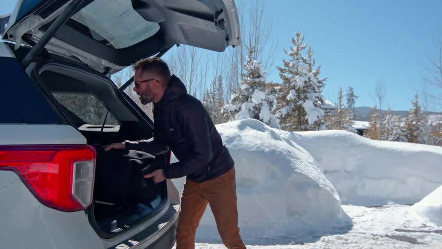 A man walks out of a garage holding a guitar and places it in the trunk of a car parked outside surrounded by snow at midday