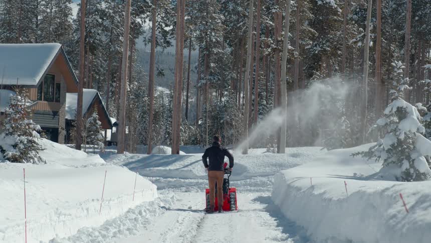 A man operates an orange snow blower to clear a path through a deep snowcovered landscape with pine trees and houses in the background on a sunny winter day