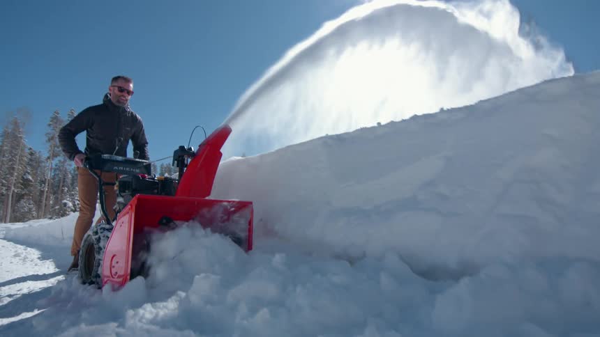 A man wearing sunglasses and a black jacket using a snow plow to clear snow from the road at midday