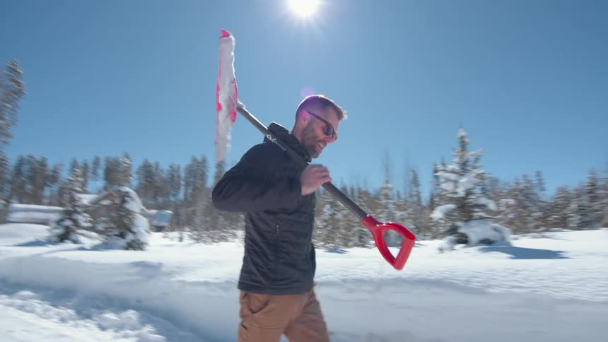 Man walks in the snow carrying a red shovel on his shoulder