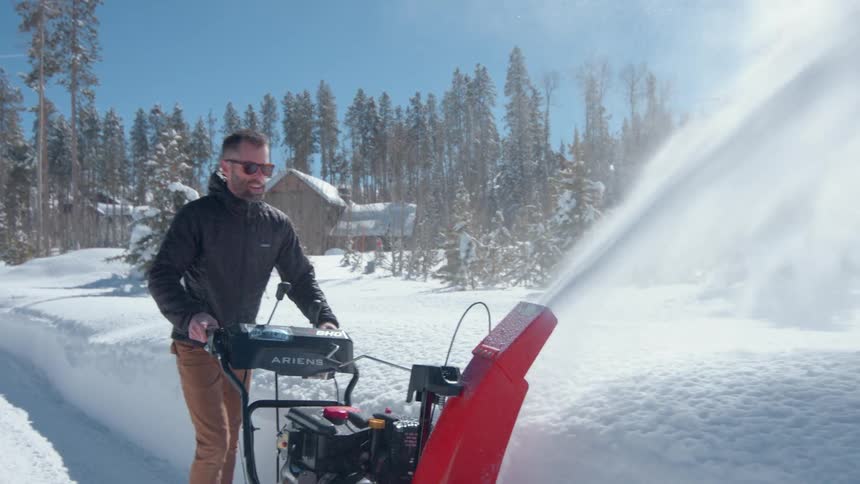 A man in sunglasses clears snow with a red ariens snowblower under bright sun and blue skies