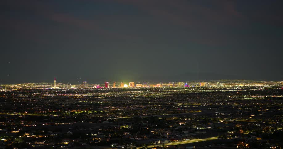 The video shows an aerial shot of a cityscape at night with a vast expanse of lights stretching out towards a distant horizon in the distance a group of brightly lit taller buildings form a distinct skyline