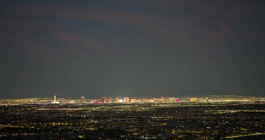A distant panoramic view of a vast illuminated city skyline at night with a brightly lit strip of buildings in the foreground against a dark and cloudy sky