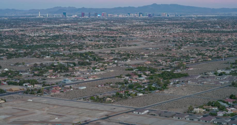 A desert suburb stretches wide with scattered houses empty plots and sparse vegetation under a hazy sky