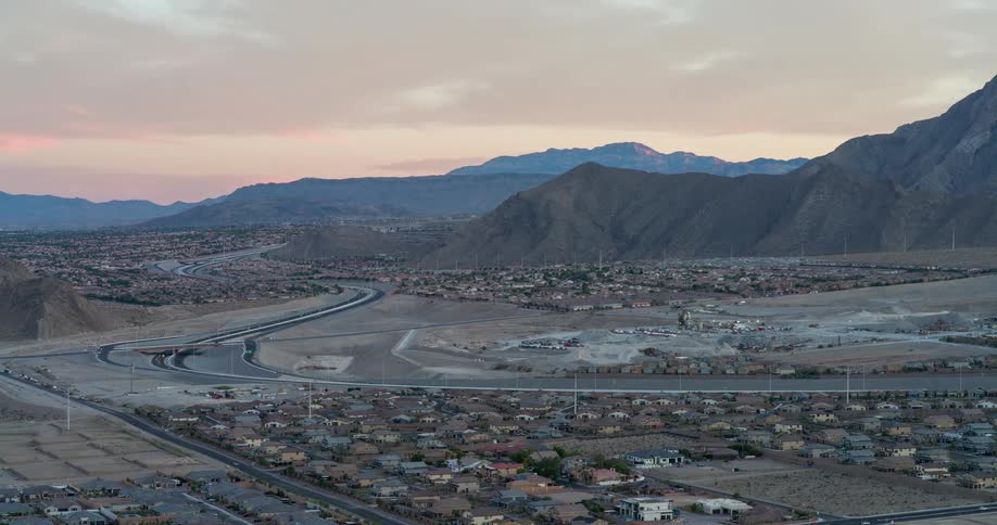 The video shows a scenic aerial shot of a suburban neighborhood situated in a valley the houses are nestled at the base of large jagged mountains with a highway curving through the landscape