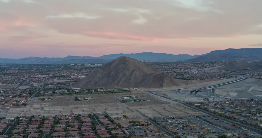 An aerial view of a small mountain or large hill rising from the middle of a developing suburban area with houses and paved streets surrounding it all set against a backdrop of distant mountains and a hazy colorful sunset sky